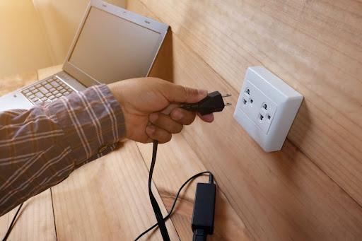 A close-up of a hand plugging a laptop charger into an outlet.